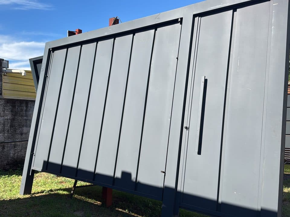 Portão de aço cinza com frisos verticais largos e portão social com puxador longo, pronto no pátio