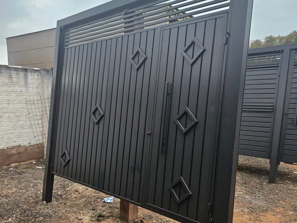 Portão de aço preto com frisos verticais, ripas no topo e dois losangos, pronto no pátio